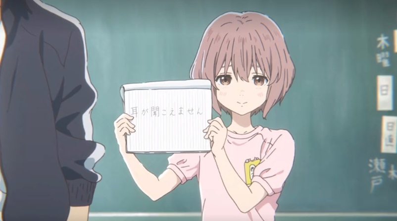 Uma garota de cabelo curto castanho segurando um caderno com seu nome escrito em japonês, está usando uma blusinha rosa.
