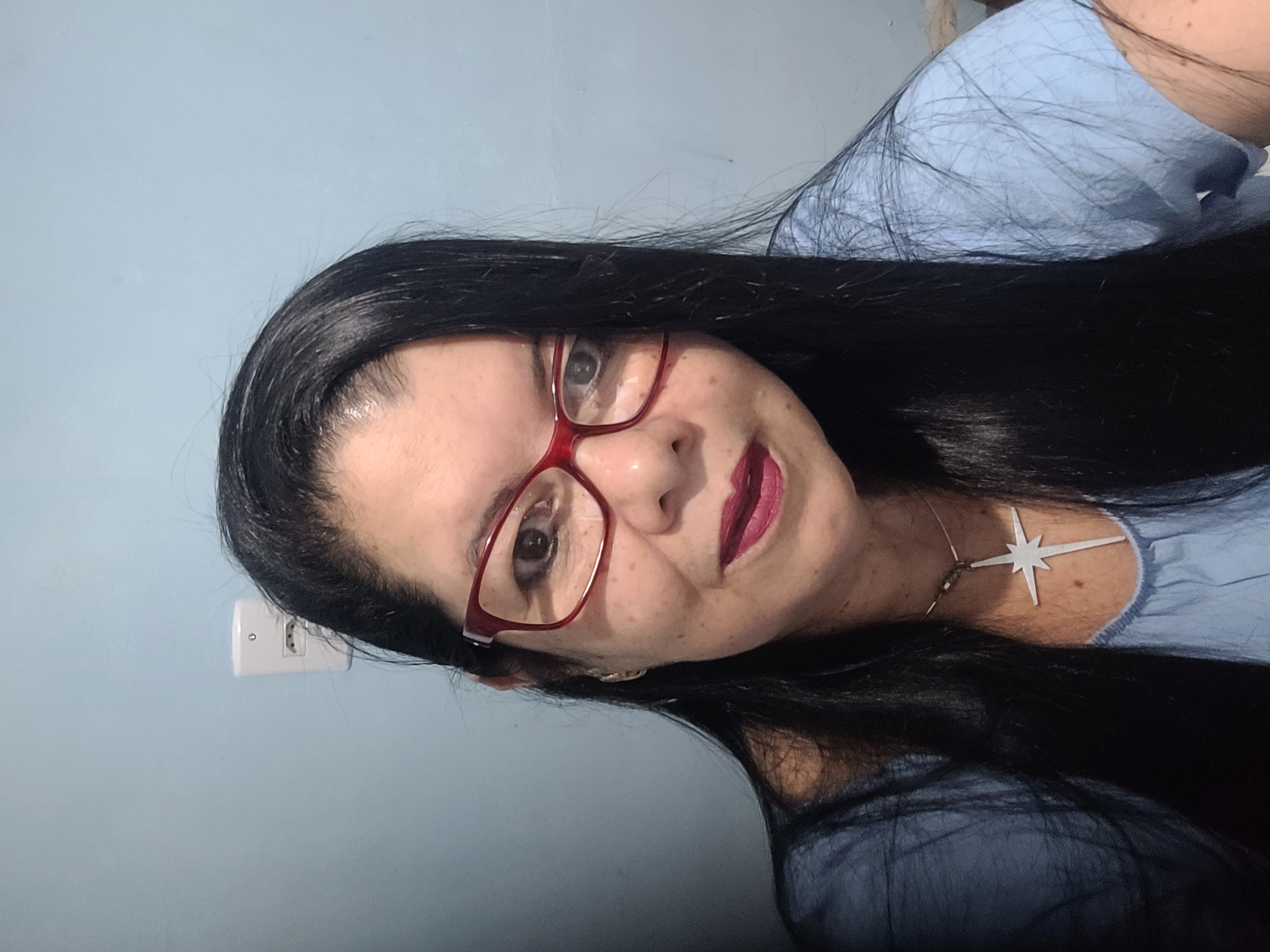 A imagem é uma foto da autora do curso. Pele branca, cabelos pretos longos, usando um óculos de armação na cor vermelha.